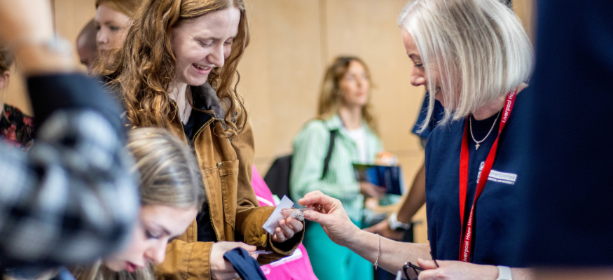A student taking a badge from a staff member at a busy event, she is smiling and looking down at her hands as she takes it